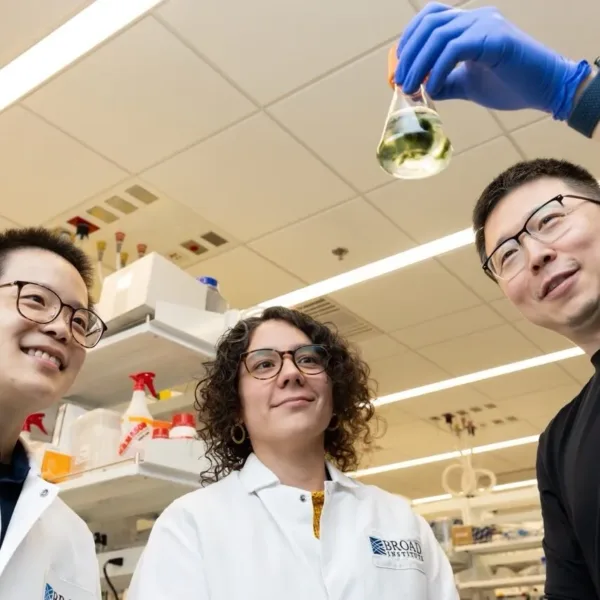 Feng Zhang, the co-director of the Molecular Therapeutics center is holding up an Erlenmeyer flask with some liquid and floating biological material in it while standing next to two researchers in his lab.