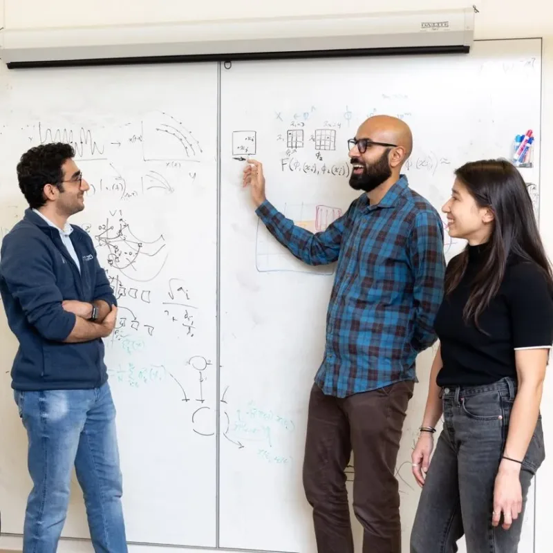 Three researchers: two male and one female, stand in front of a white board with dry erase markers discussing computational equations
